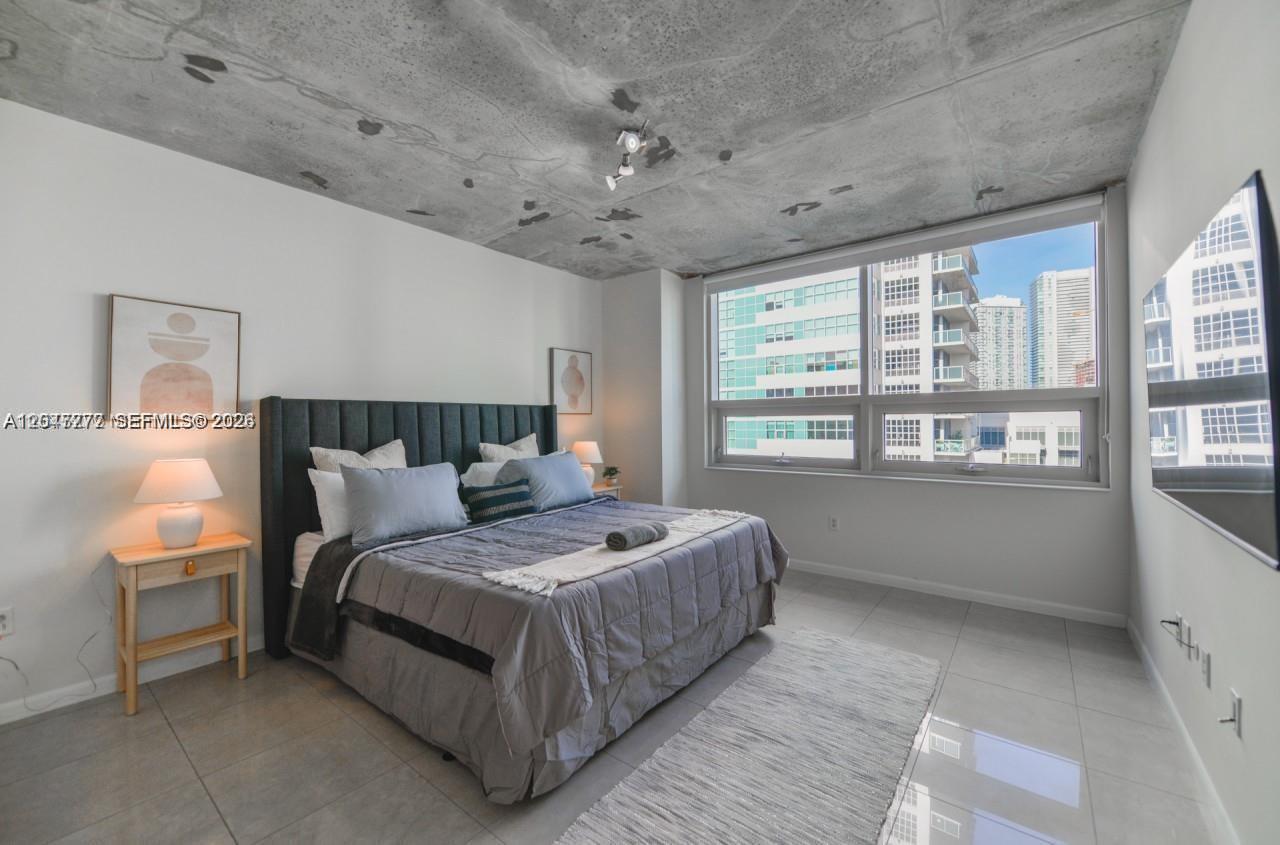 A bedroom features a gray bed, concrete ceiling, and large windows overlooking city buildings — 3250 NE 1st Ave # 901, Miami FL 33137, MIAMI, FL