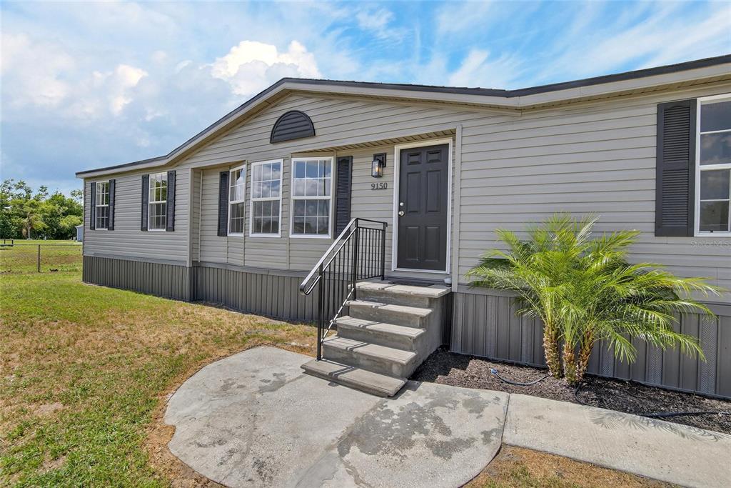 A gray manufactured home features a dark front door, concrete steps, and a small palm — 9150 WOOD TERRACE DRIVE, TAMPA, FL