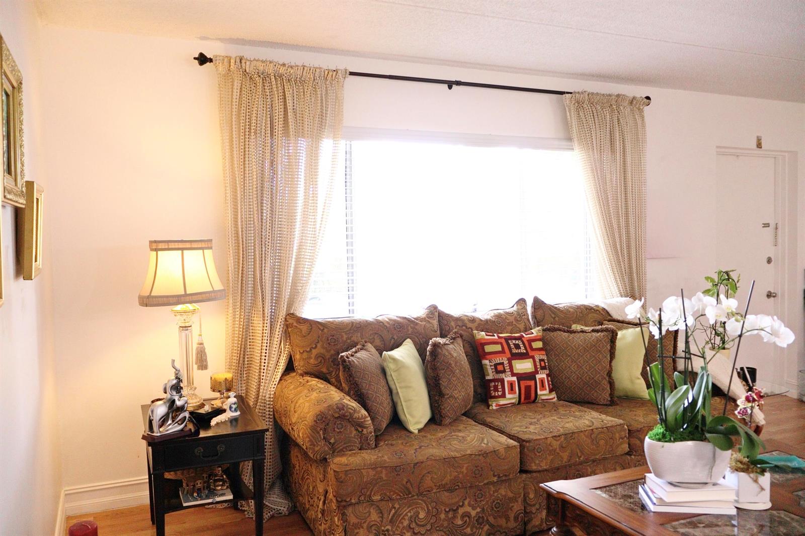 A living room features a patterned sofa, a side table with a lamp, and curtains — 3400 Springdale Boulevard # 214, Palm Springs FL 33461, PALM SPRINGS, FL