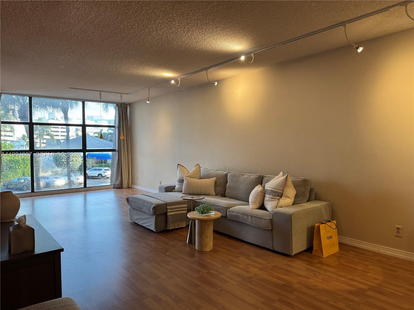 A living room features a gray sectional sofa, wood flooring, track lighting, and floor-to-ceiling windows — 600 Parkview Dr # 222, Hallandale Beach FL 33009, HALLANDALE BEACH, FL