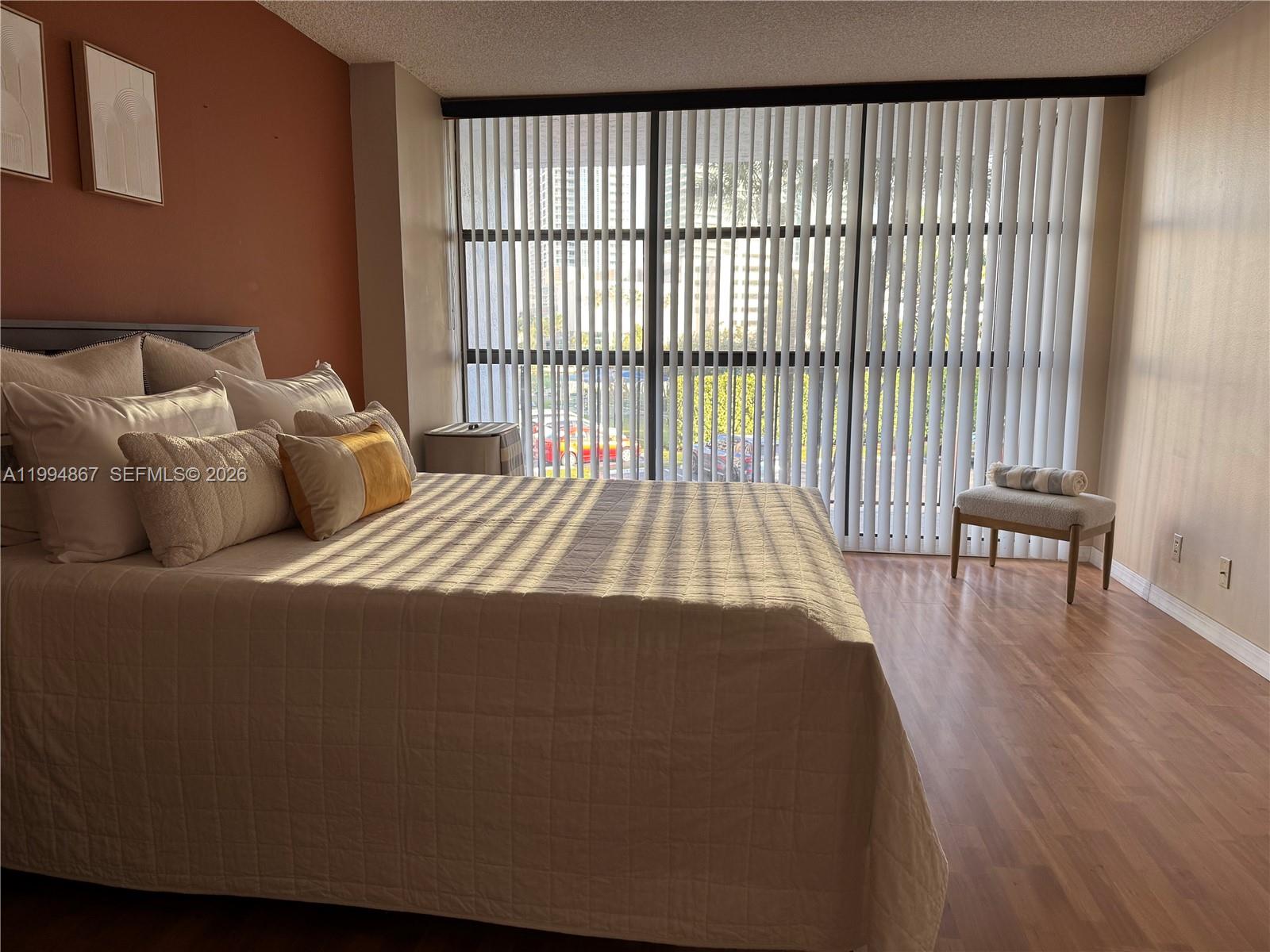 A bedroom features a bed with neutral bedding, wood-look flooring, and floor-to-ceiling vertical blinds — 600 Parkview Dr # 222, Hallandale Beach FL 33009, HALLANDALE BEACH, FL