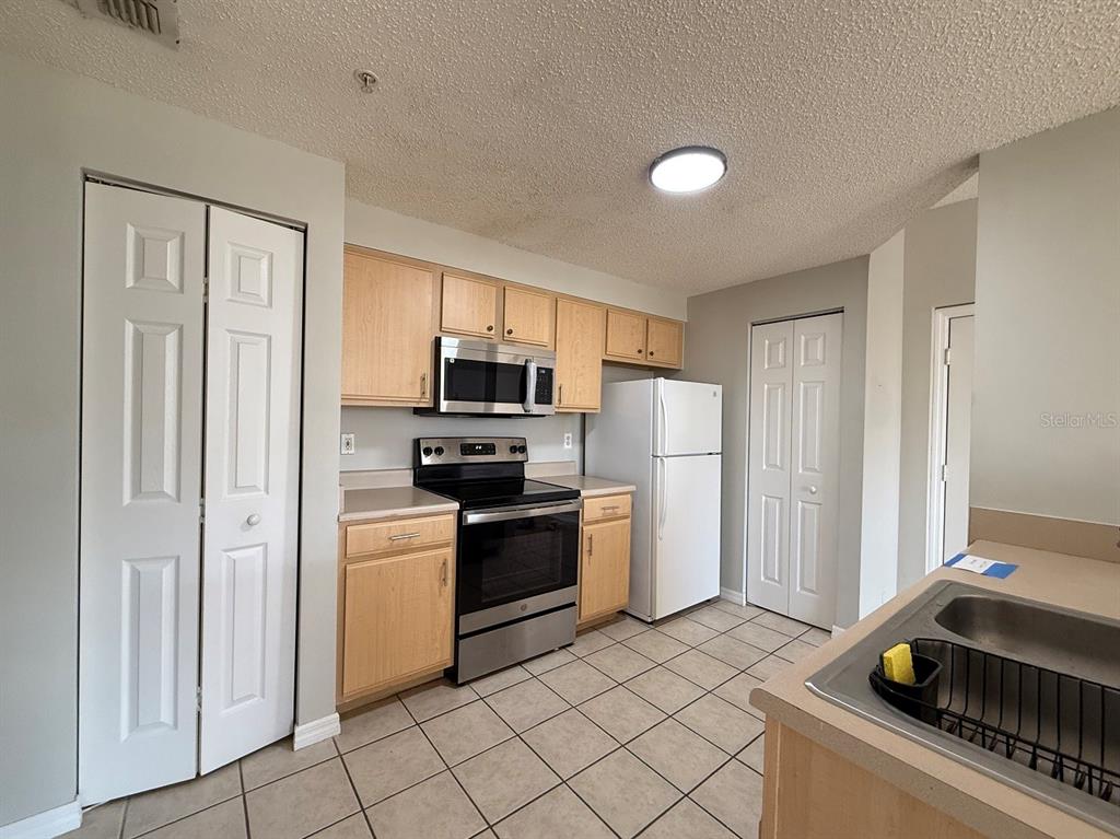 A kitchen features light wood cabinets, stainless steel appliances, a white refrigerator, and tile flooring — 10831 WINDSOR WALK DRIVE UNIT 1305, ORLANDO, FL