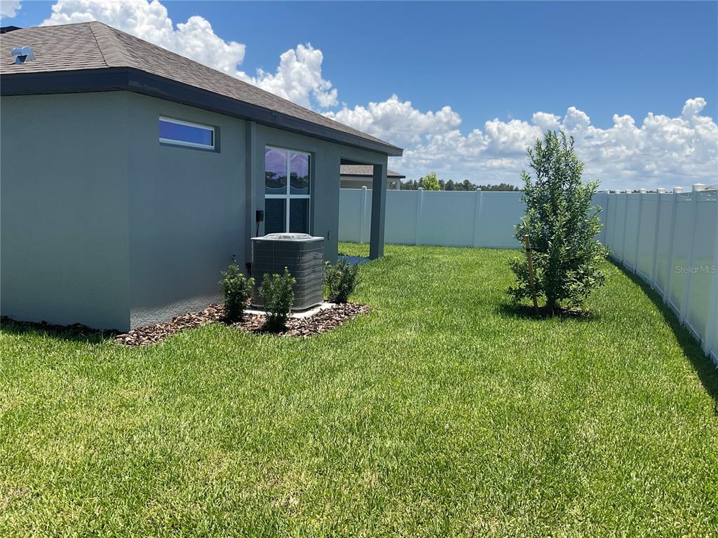 A gray house exterior features a backyard lawn, an HVAC unit, and a white fence — 3517 HORNBEAM ROAD, SPRING HILL, FL
