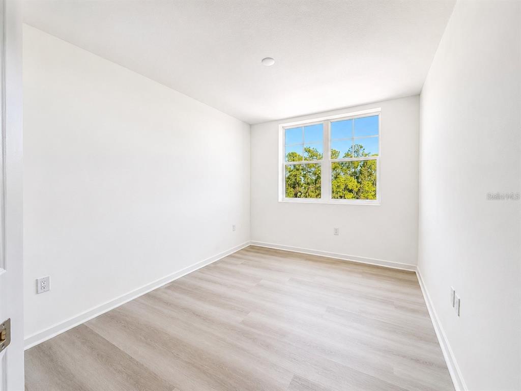 An empty bedroom features white walls, light wood-look flooring, and a window overlooking trees — 5587 MILLENIA PARK BOULEVARD UNIT 316, ORLANDO, FL