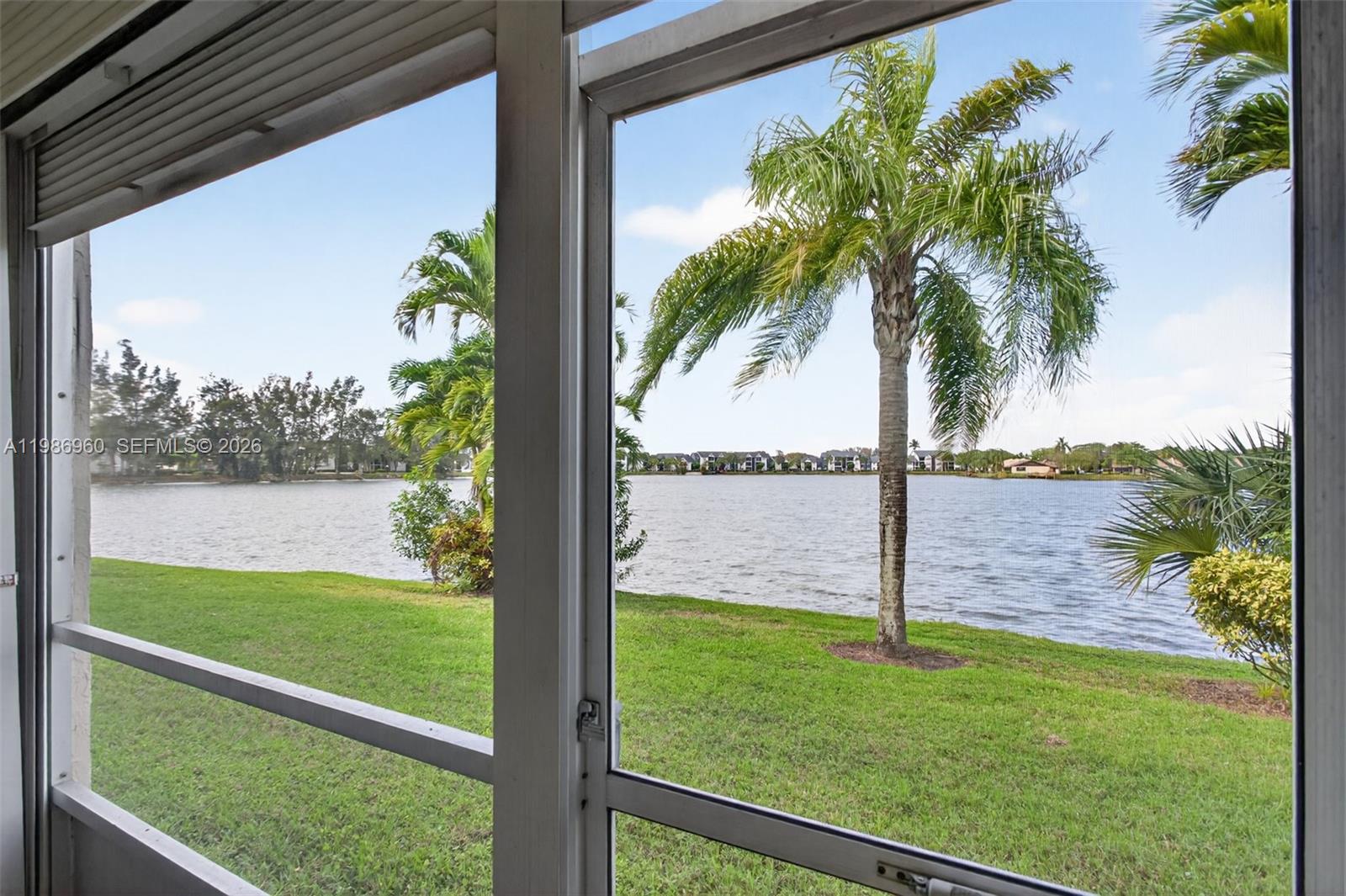 A view of a lake and palm trees seen through a screened-in porch window — 5520 Lakeside Dr # 105, Margate FL 33063, MARGATE, FL