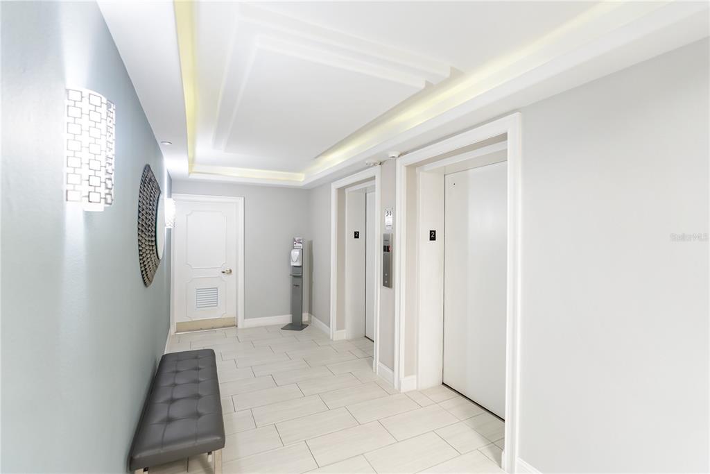 A hallway features two elevators, a white door, a grey bench, and tiled flooring — 1212 BENJAMIN FRANKLIN DRIVE UNIT 206, SARASOTA, FL
