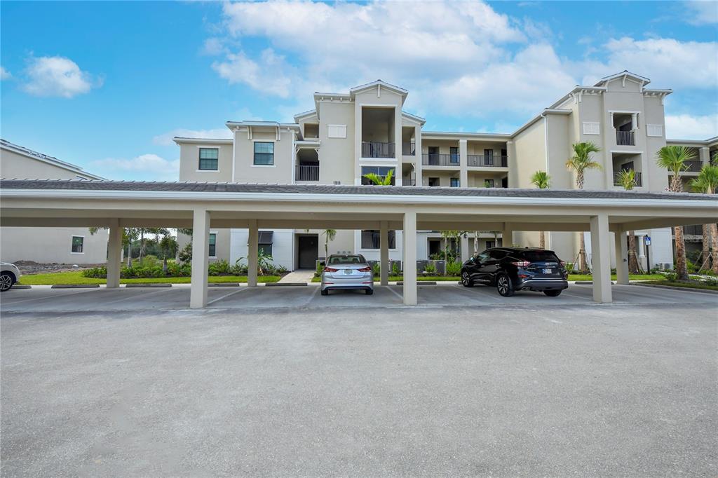 A covered parking structure sits in front of a multi-story apartment building under a blue sky — 17810 GAWTHROP STREET UNIT 101, LAKEWOOD RANCH, FL