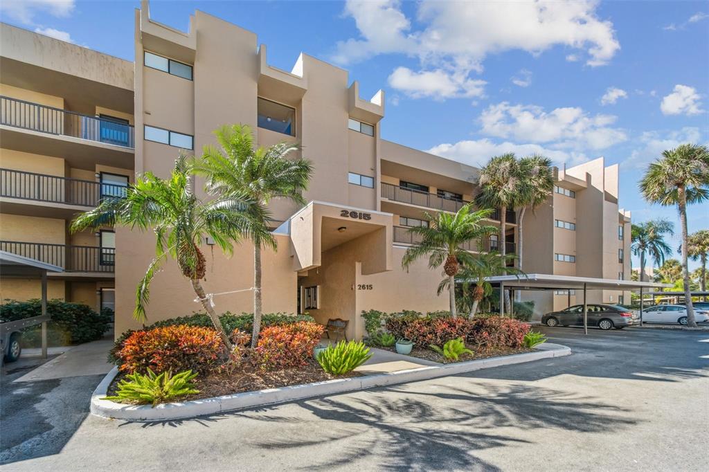 A multi-story tan apartment building features a covered entrance, palm trees, and parking spaces — 2615 COVE CAY DRIVE UNIT 409, CLEARWATER, FL
