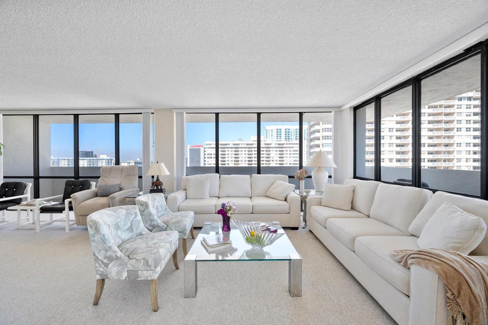 A bright living room features white sofas, patterned armchairs, a glass coffee table, and floor-to-ceiling windows — 2017 S Ocean Drive, Hallandale Beach FL 33009, HALLANDALE BEACH, FL