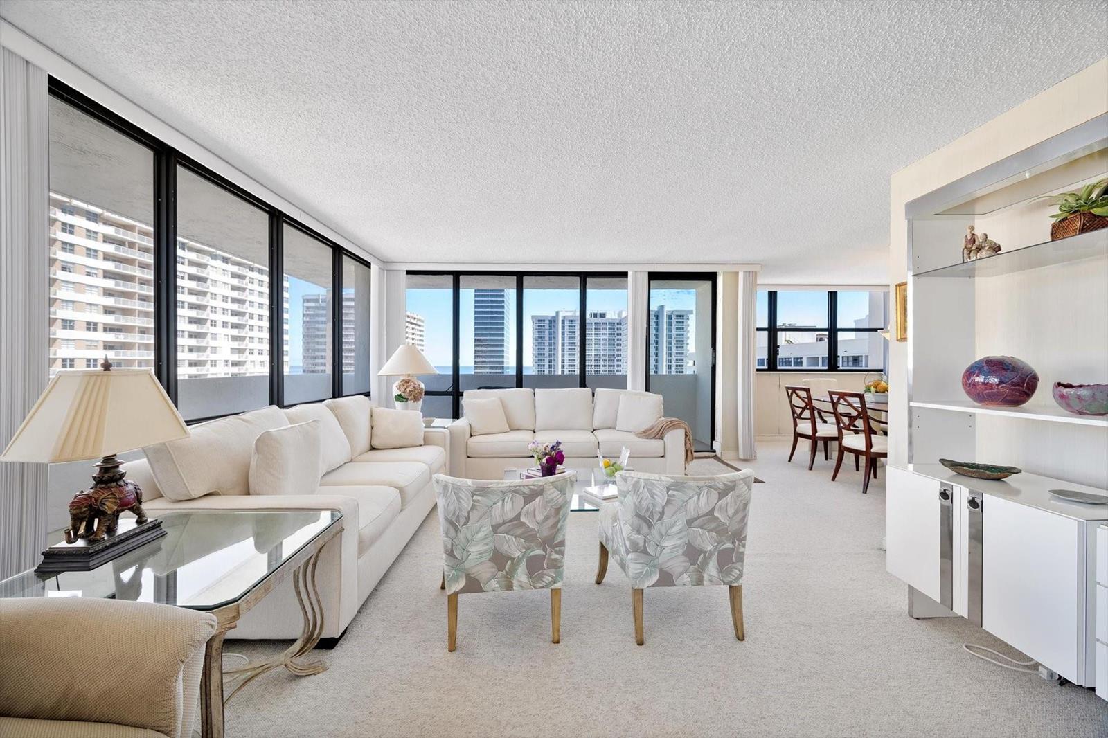 A bright living room features white sofas, patterned armchairs, and floor-to-ceiling windows overlooking city buildings — 2017 S Ocean Drive, Hallandale Beach FL 33009, HALLANDALE BEACH, FL