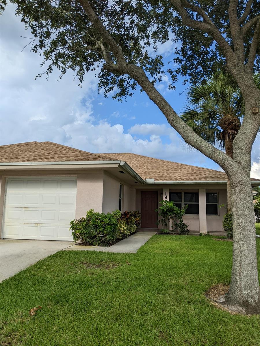 A single-story house with a white garage door, tan stucco walls, and a large tree — 1804 Blue Heron Lane # C, Fort Pierce FL 34982, FORT PIERCE, FL
