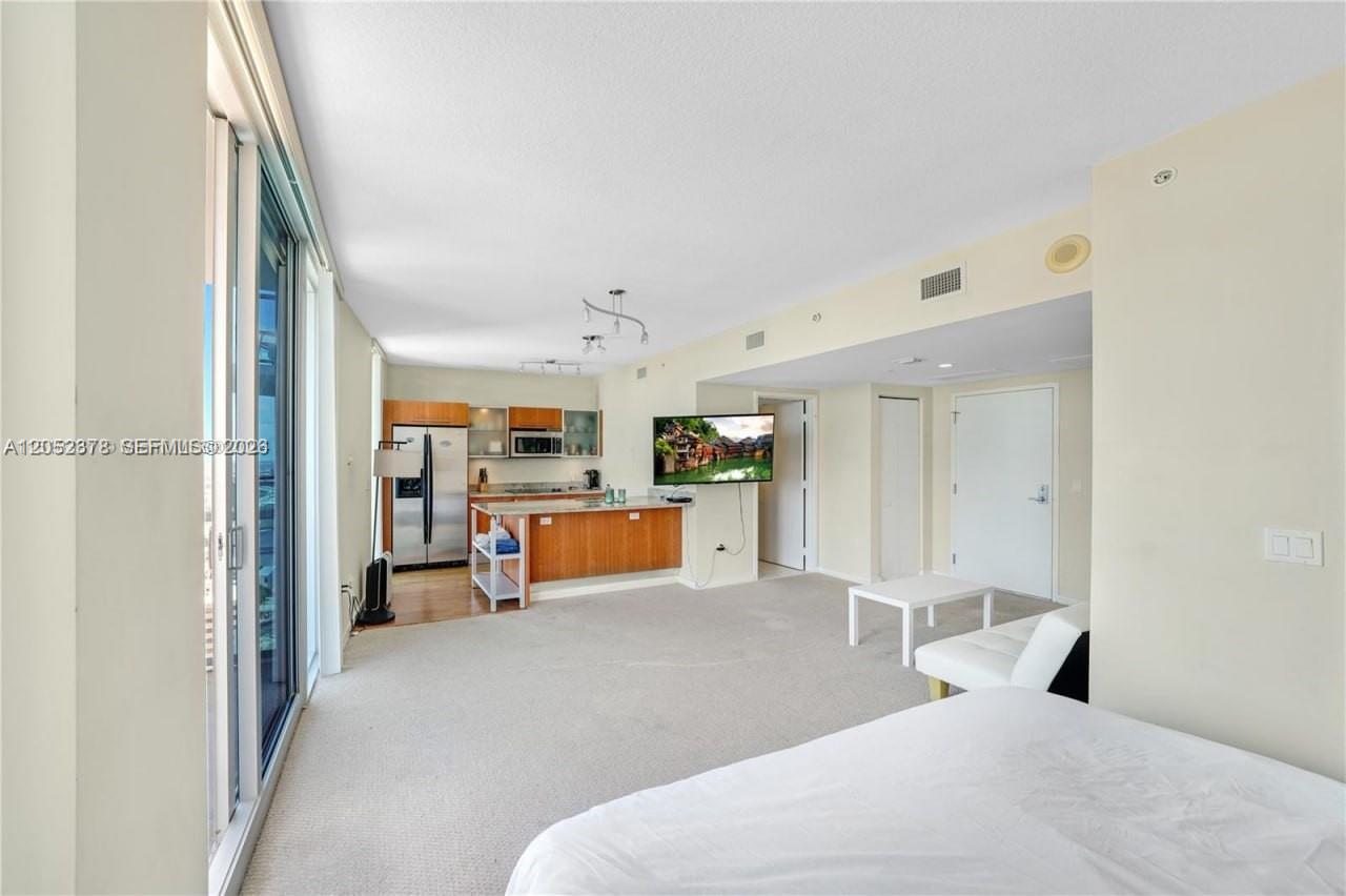 A living room with beige carpet features an open kitchen with wood cabinets and appliances — 185 SW 7th St # 3100, Miami FL 33130, MIAMI, FL