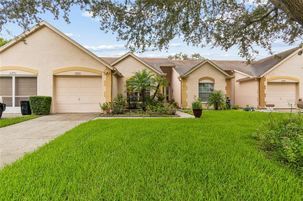 A single-story duplex with beige stucco walls, two garage doors, and a front lawn — 11512 VERSAILLES LANE UNIT 11512, PORT RICHEY, FL