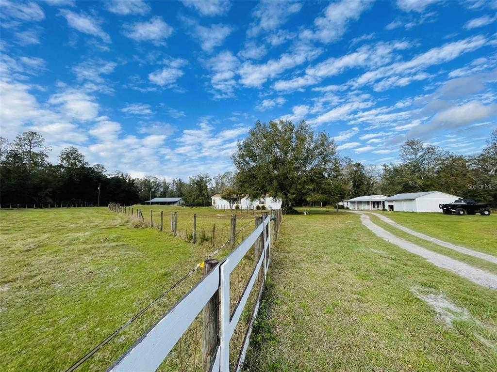 A white fence borders a grassy field leading toward a house and detached garage — 6810 PEARSON LANE, WESLEY CHAPEL, FL