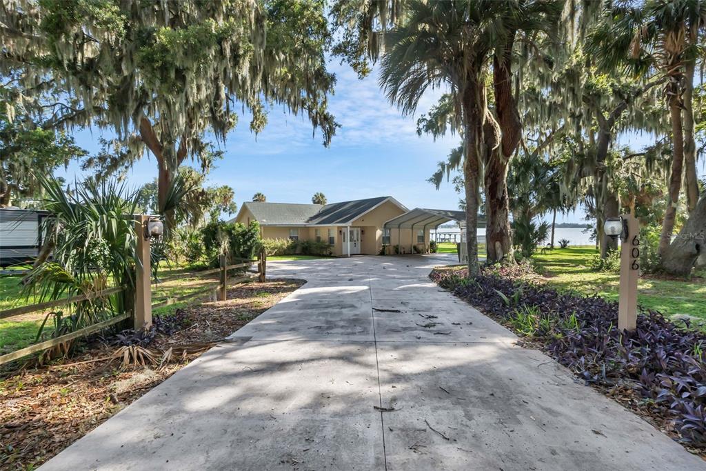 A concrete driveway leads to a single-story house with a carport under large oak trees — 606 N SHORE DRIVE, LEESBURG, FL