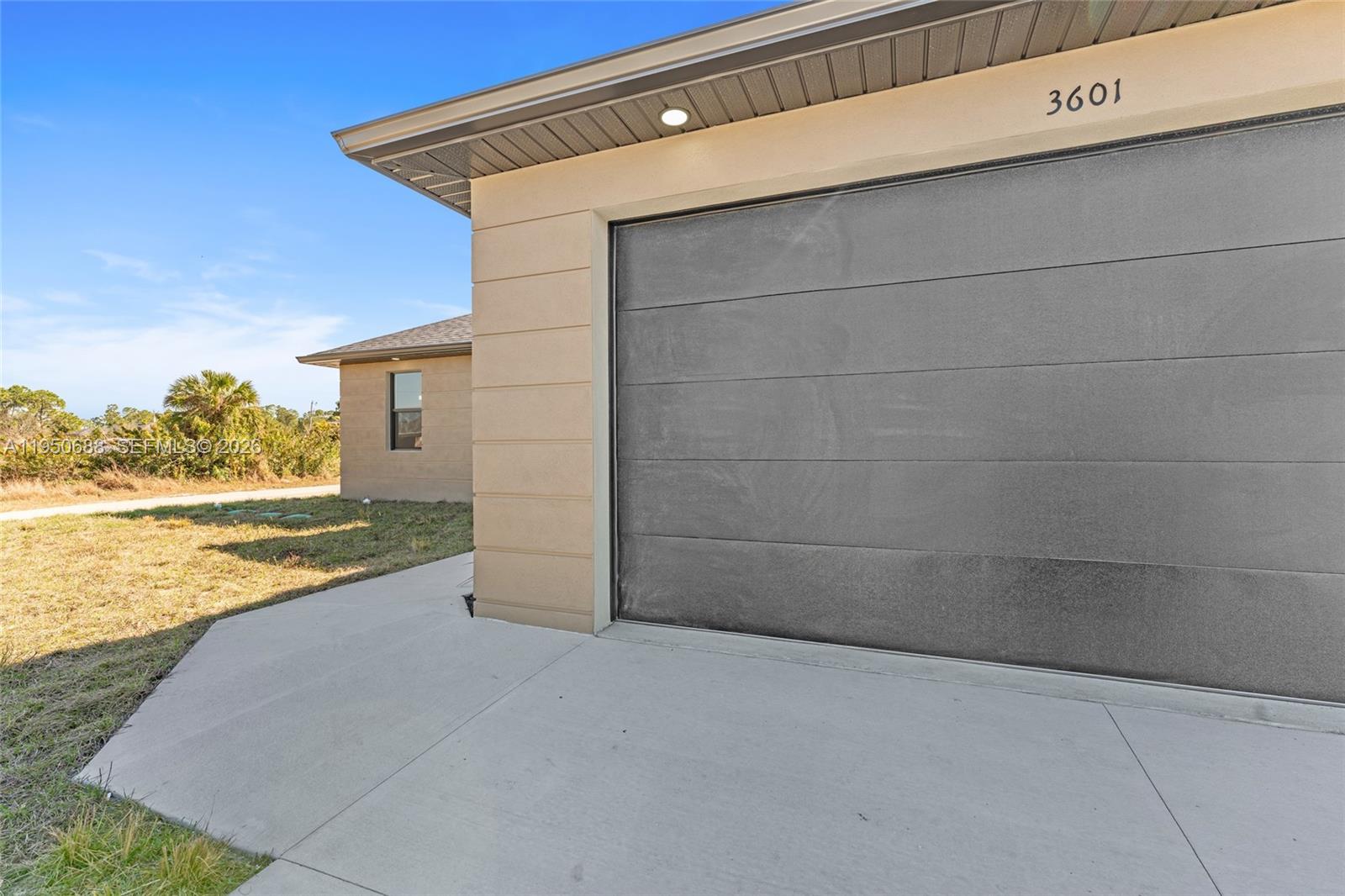 A concrete driveway leads to a gray garage door on a tan stucco house — 3601 28th St SW, Lehigh Acres FL 33976, LEHIGH ACRES, FL