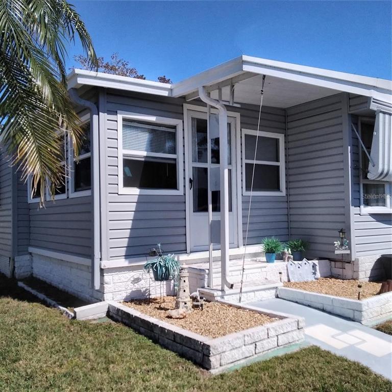A gray mobile home exterior features a white front door, small porch, and stone planters — 10473 HOLIDAY SHORES DRIVE UNIT 303, LARGO, FL