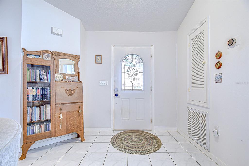 A tiled entryway features a wooden bookshelf, a white front door, and a wall-mounted thermostat — 6911 OAKCREST WAY, ZEPHYRHILLS, FL