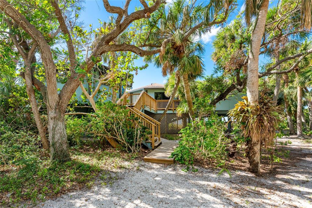 An elevated house with a wooden staircase and deck surrounded by trees and sandy ground — 6580 MANASOTA KEY ROAD, ENGLEWOOD, FL
