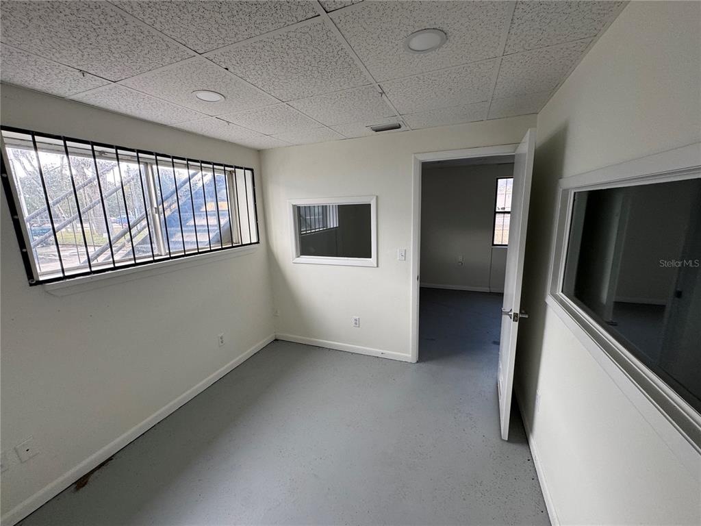 An office room with gray flooring, white walls, a barred window, and an interior doorway — 702 W PARK AVENUE UNIT A1, EDGEWATER, FL