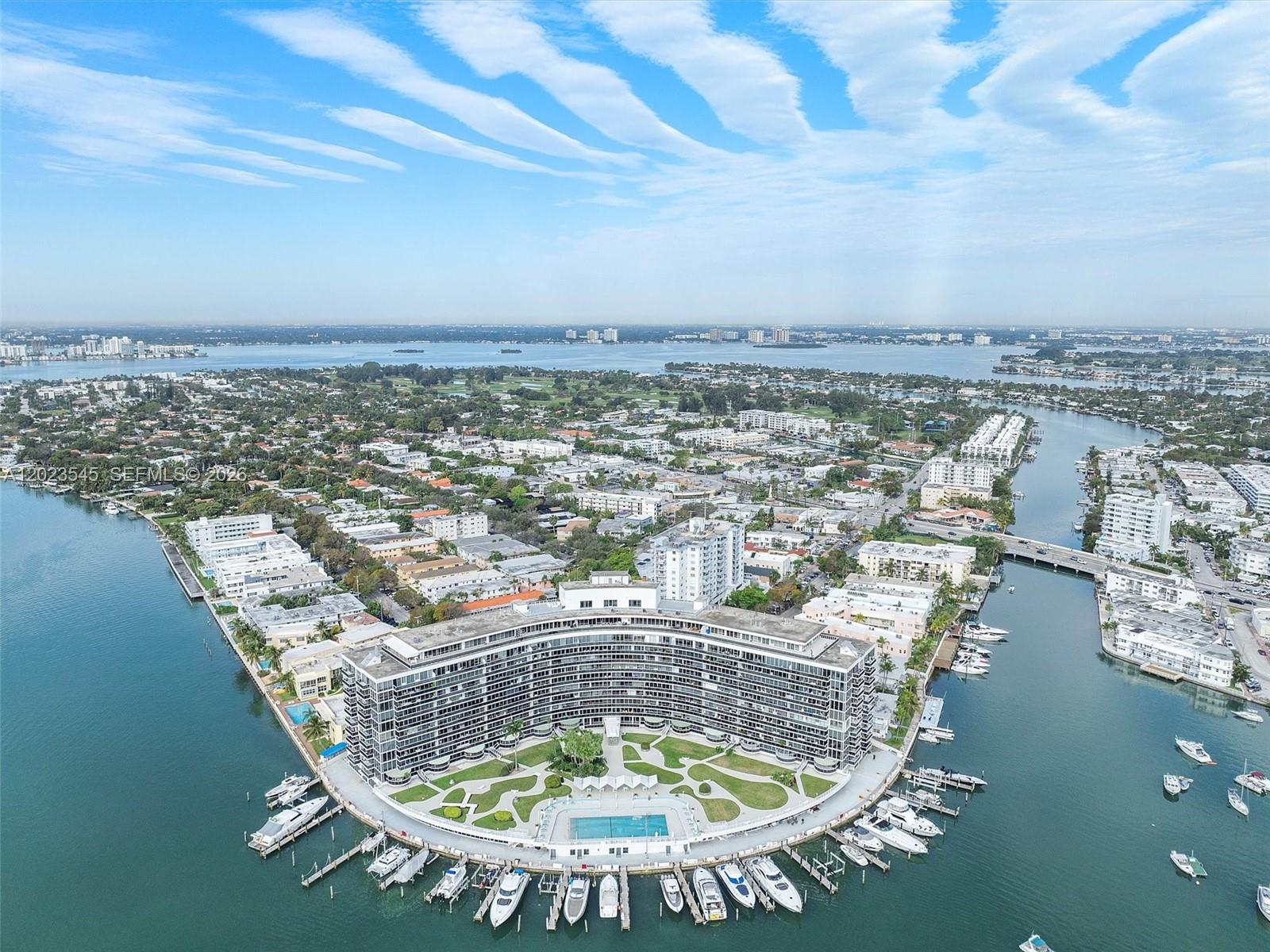 An aerial view shows a curved waterfront apartment building with a pool and boat docks — 900 Bay Dr # 1027, Miami Beach FL 33141, MIAMI BEACH, FL