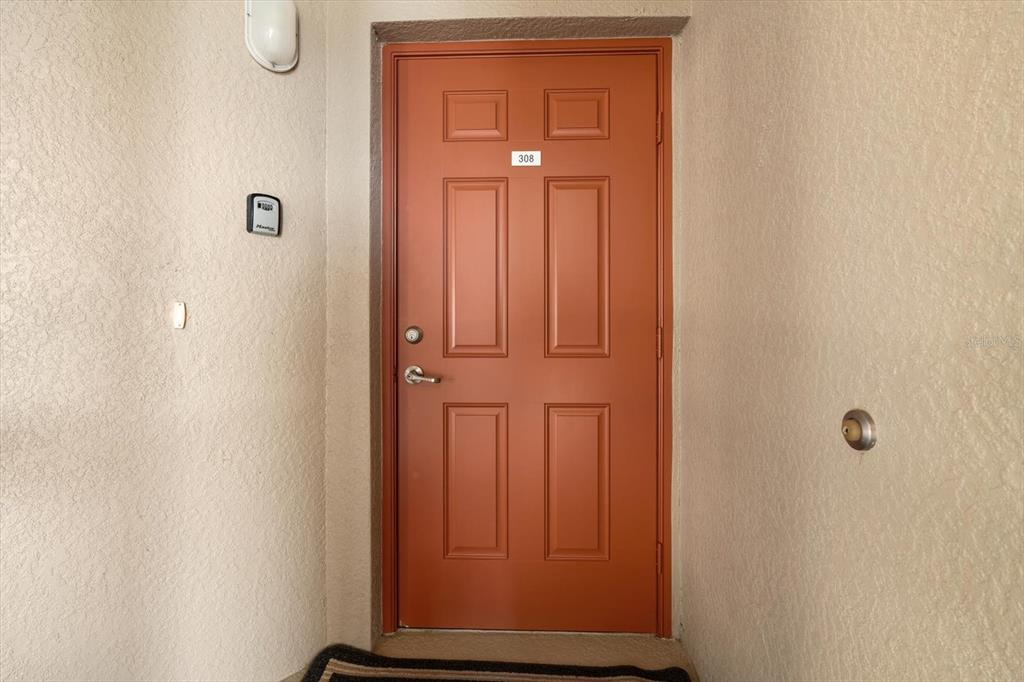 A red front door with the number 308 is set into a beige stucco wall — 7803 GRAND ESTUARY TRAIL UNIT 308, BRADENTON, FL