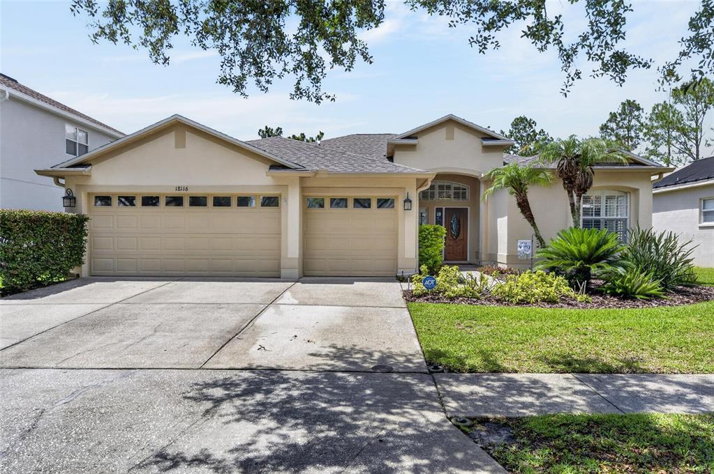 A single-story house with a beige stucco exterior, a three-car garage, and a landscaped yard — 18116 KARA CT, TAMPA, FL