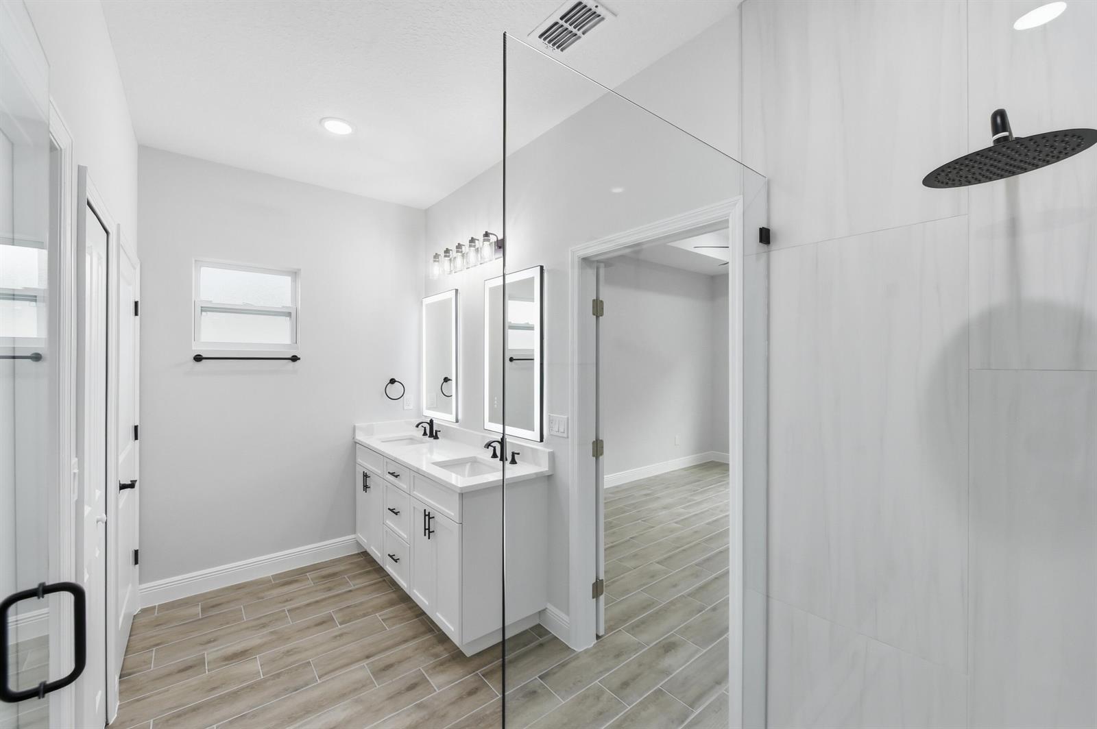A modern bathroom features a double vanity, a glass-enclosed shower, and wood-look tile flooring — 7825 96th Avenue, Vero Beach FL 32967, VERO BEACH, FL