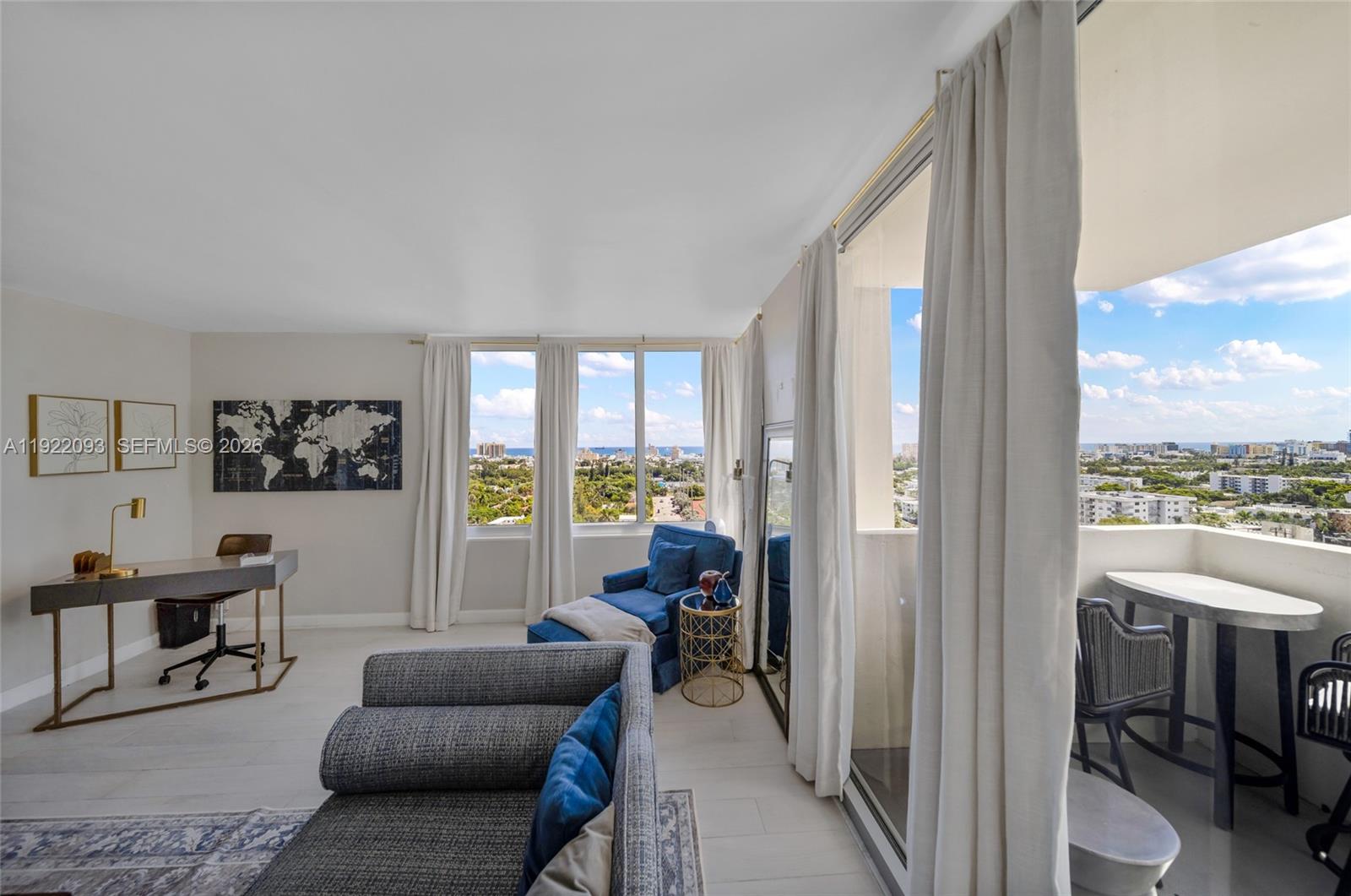 A living room with a desk, blue armchair, and balcony overlooking a city and ocean — 1200 West Ave # 15, Miami Beach FL 33139, MIAMI BEACH, FL