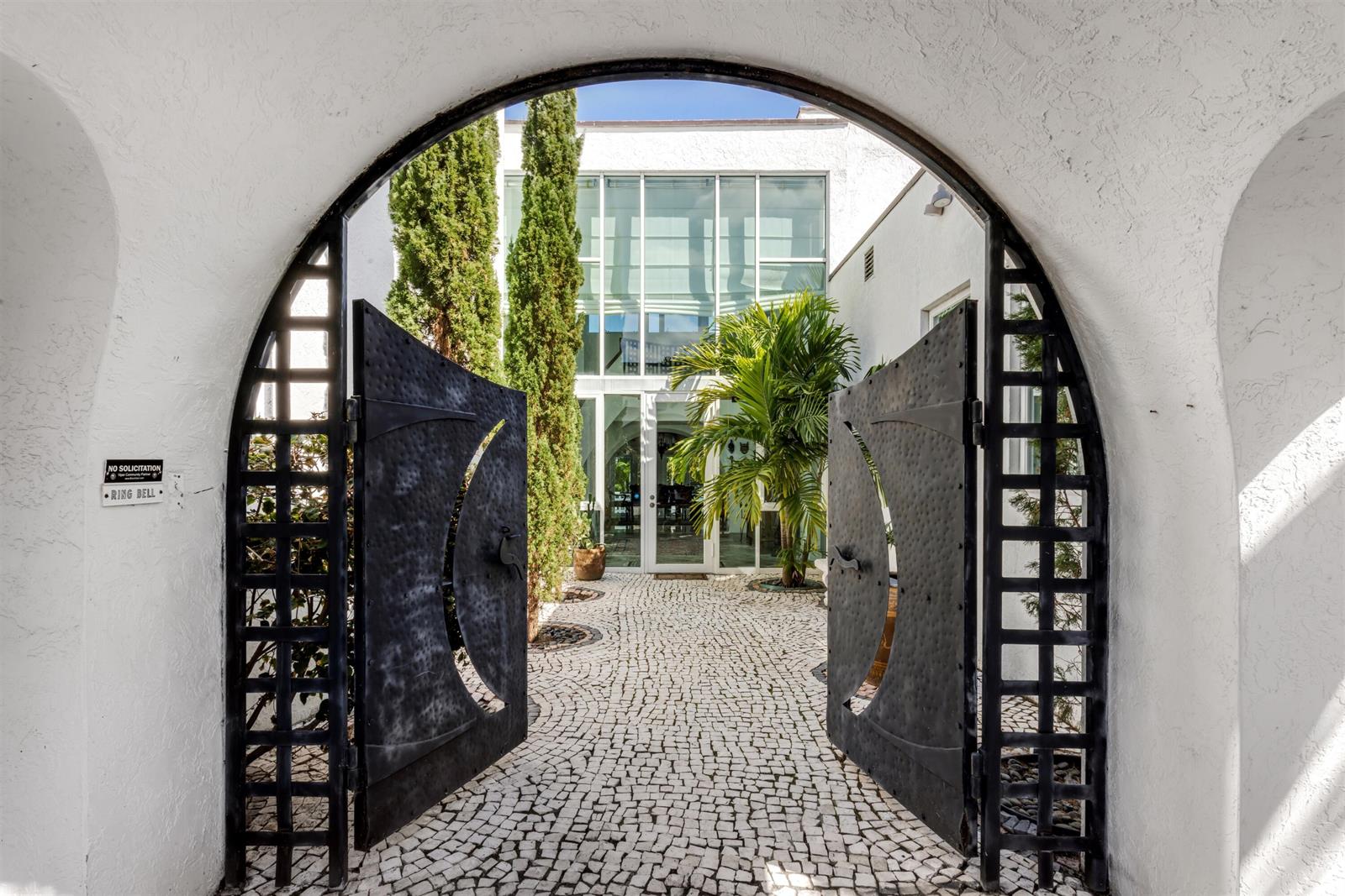 An arched gateway with black metal doors opens to a cobblestone courtyard and glass facade — 794 Staghorn Court, Boca Raton FL 33432, BOCA RATON, FL