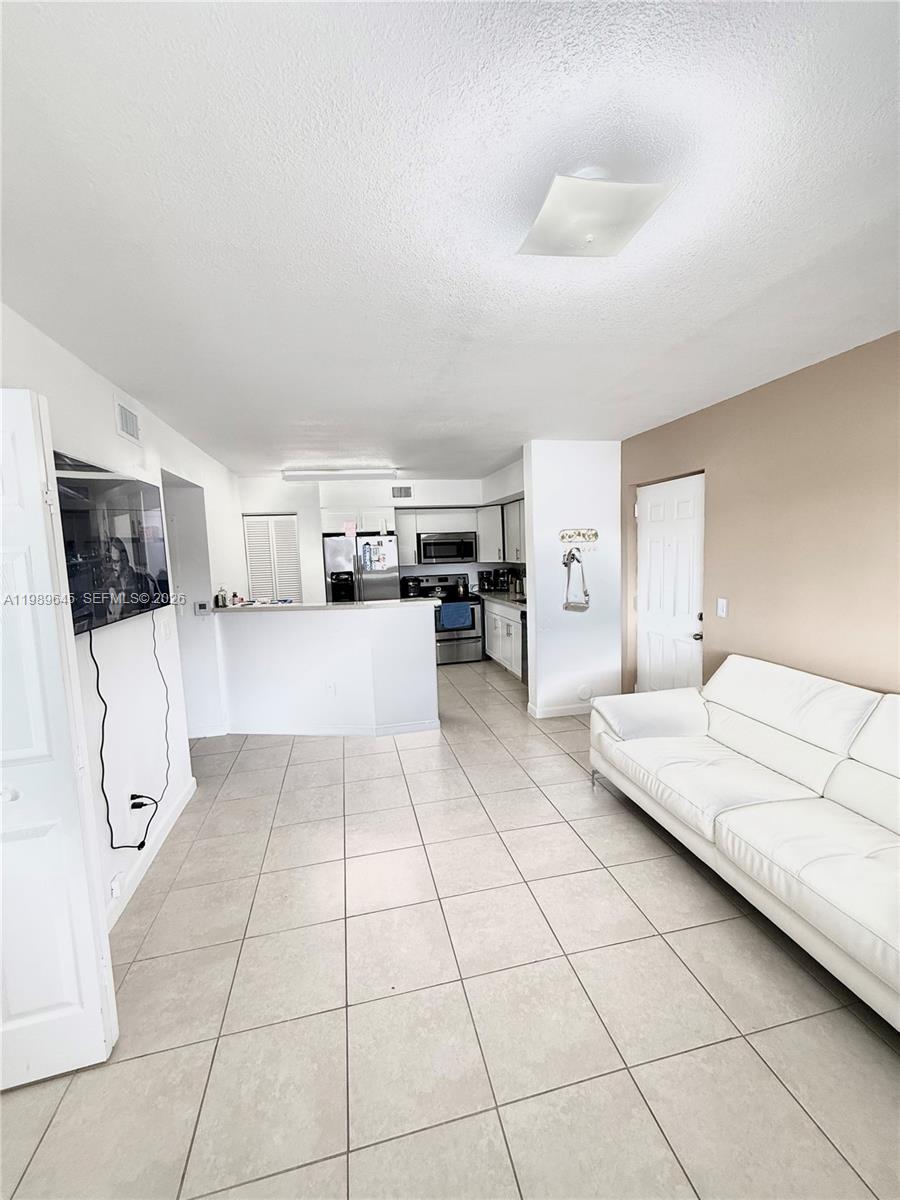 A living room with tile floors features a white sofa, a wall-mounted television, and an open kitchen — 3690 N 56th Ave # 902, Hollywood FL 33021, HOLLYWOOD, FL