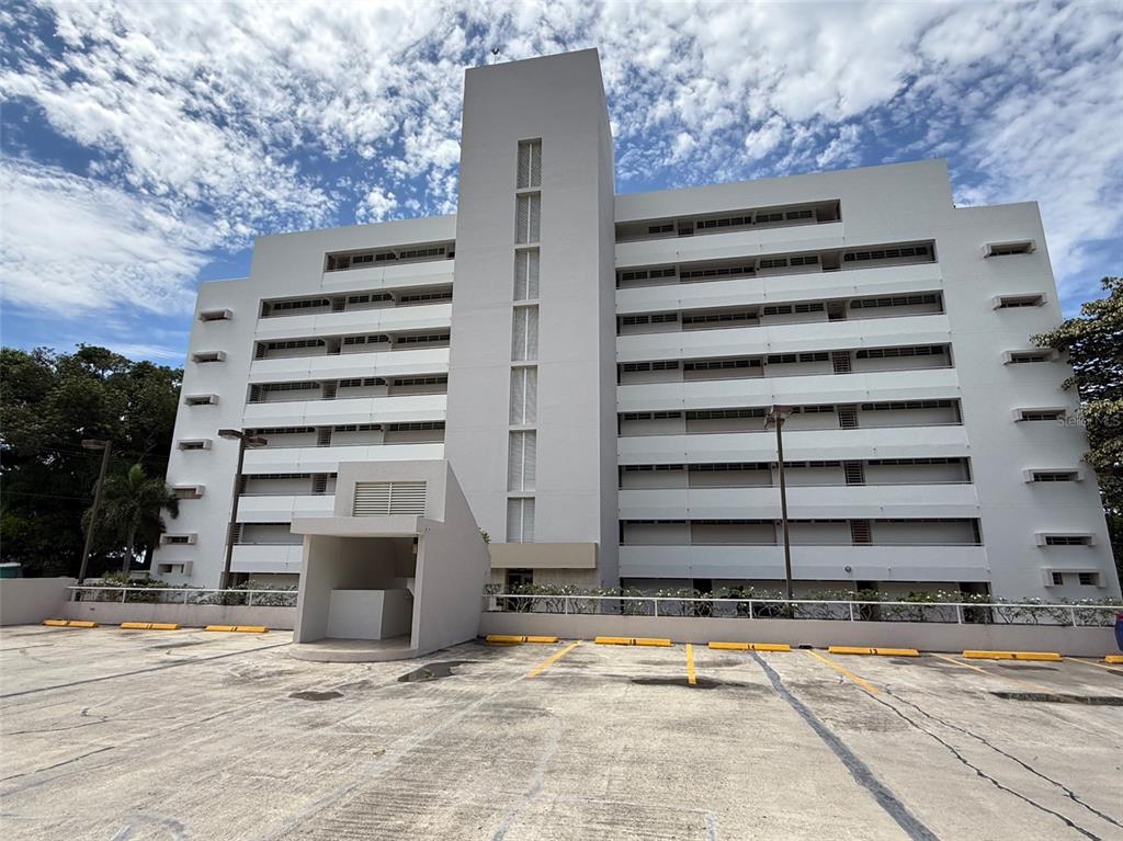 A multi-story white apartment building stands behind a paved parking lot with marked spaces — Condo Pelican Reef APT 302 UNIT 302, RINCON, PR
