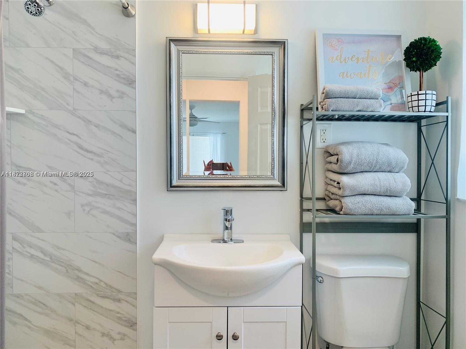 A bathroom features a white vanity, a silver-framed mirror, and a metal shelving unit — 116 SE 9th Ct, Hallandale Beach FL 33009, HALLANDALE BEACH, FL