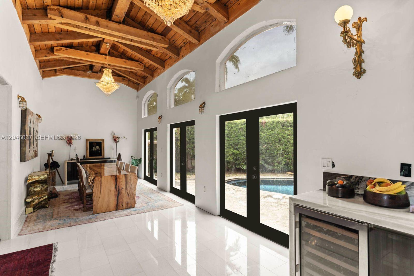 A high-ceilinged dining room features exposed wooden beams, a crystal chandelier, and glass doors overlooking a pool — 6350 Pinetree Dr, Miami Beach FL 33141, MIAMI BEACH, FL