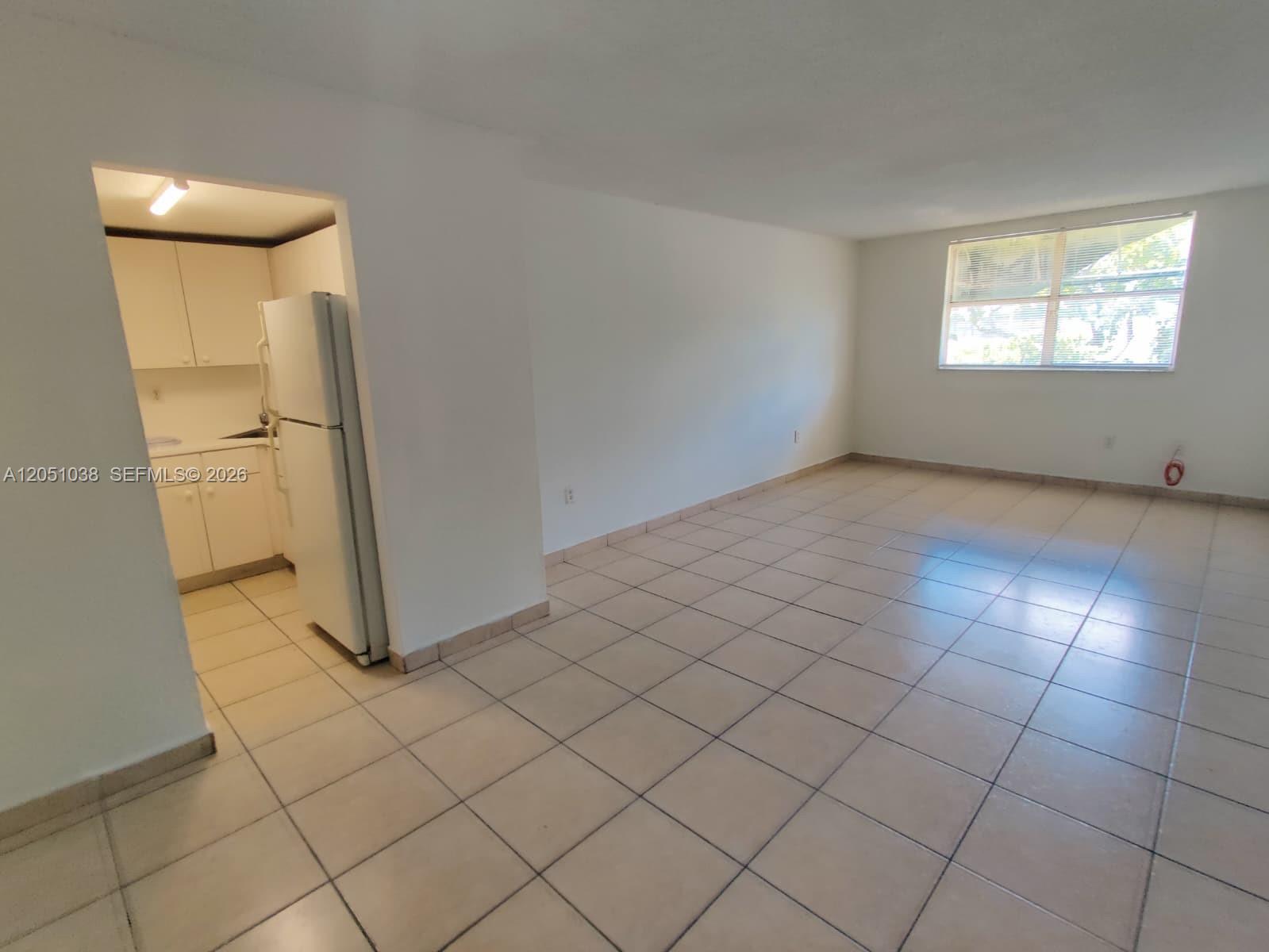 An empty room with light-colored tile flooring, white walls, and an adjacent kitchen area — 666 W 81st St # 414, Hialeah FL 33014, HIALEAH, FL