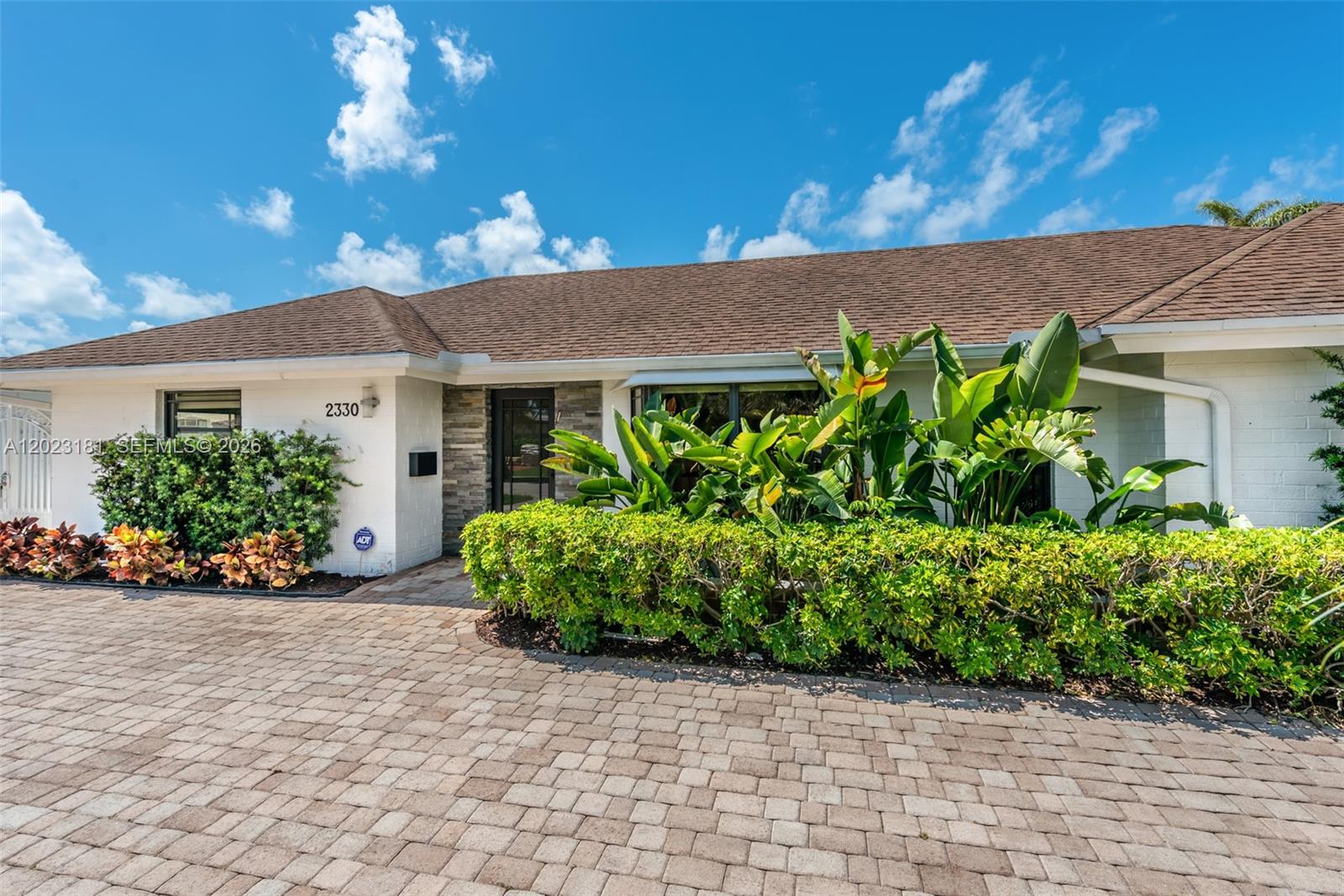 The exterior of a single-story house features a brick driveway, white walls, and lush landscaping — 2330 NE 195th St, Miami FL 33180, MIAMI, FL