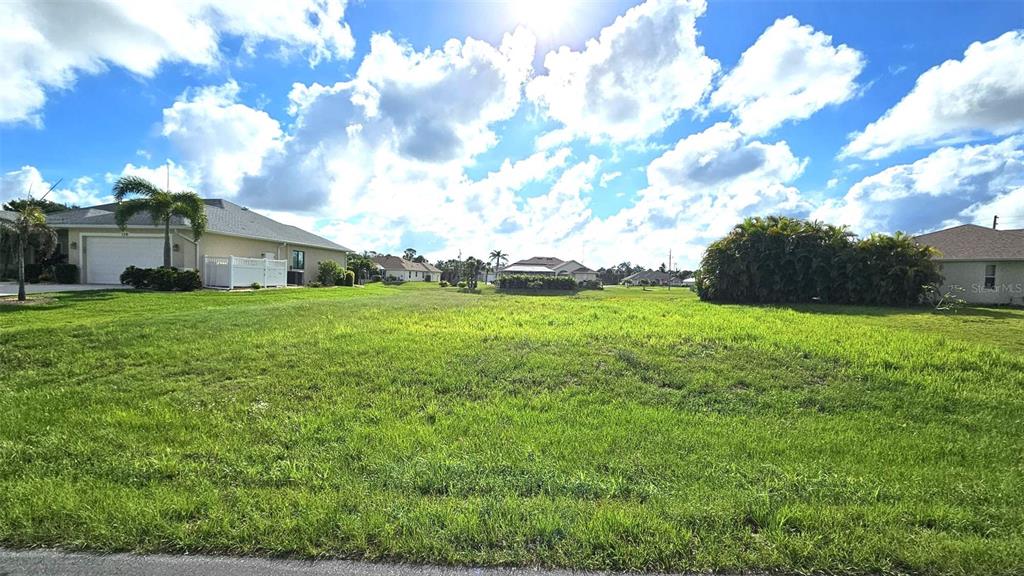 A grassy vacant lot sits between two suburban houses under a bright, cloudy sky — 144 LONG MEADOW LANE, ROTONDA WEST, FL