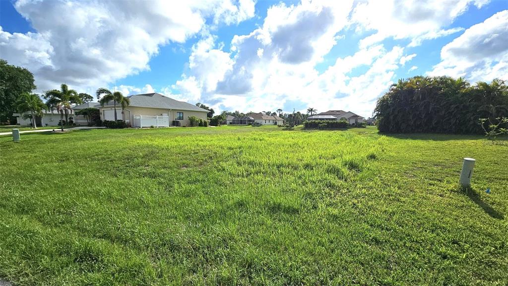 A large, grassy vacant lot is surrounded by residential houses under a cloudy blue sky — 144 LONG MEADOW LANE, ROTONDA WEST, FL