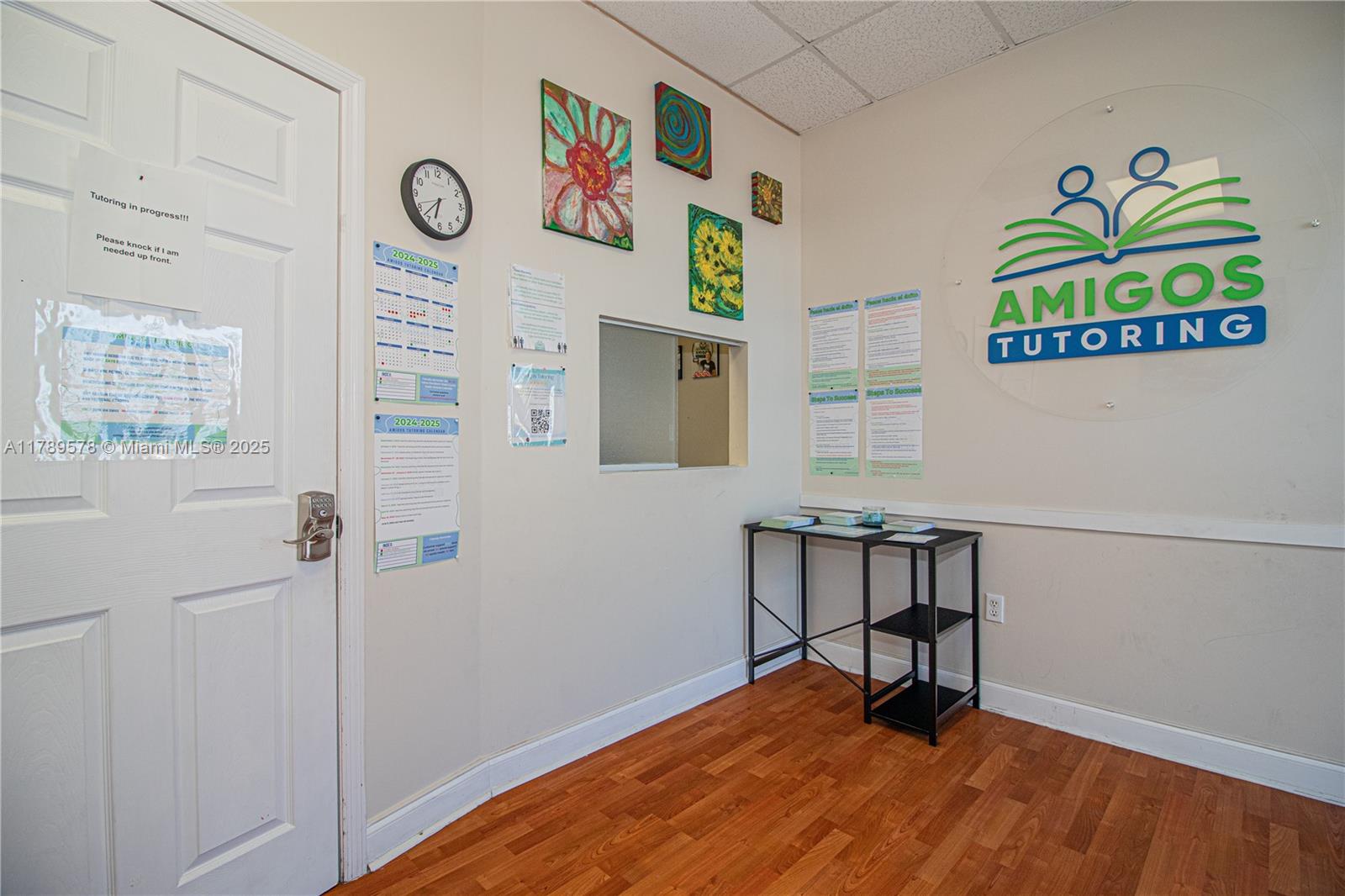 A waiting area features a white door, wood-look flooring, wall art, and an Amigos Tutoring sign — 5580 W 16th Ave, Hialeah FL 33012, HIALEAH, FL