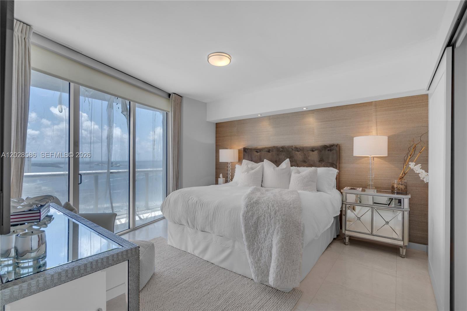 A bedroom features a bed against a wood-paneled wall, mirrored nightstands, and floor-to-ceiling windows — 465 Brickell Ave # 2703, Miami FL 33131, MIAMI, FL
