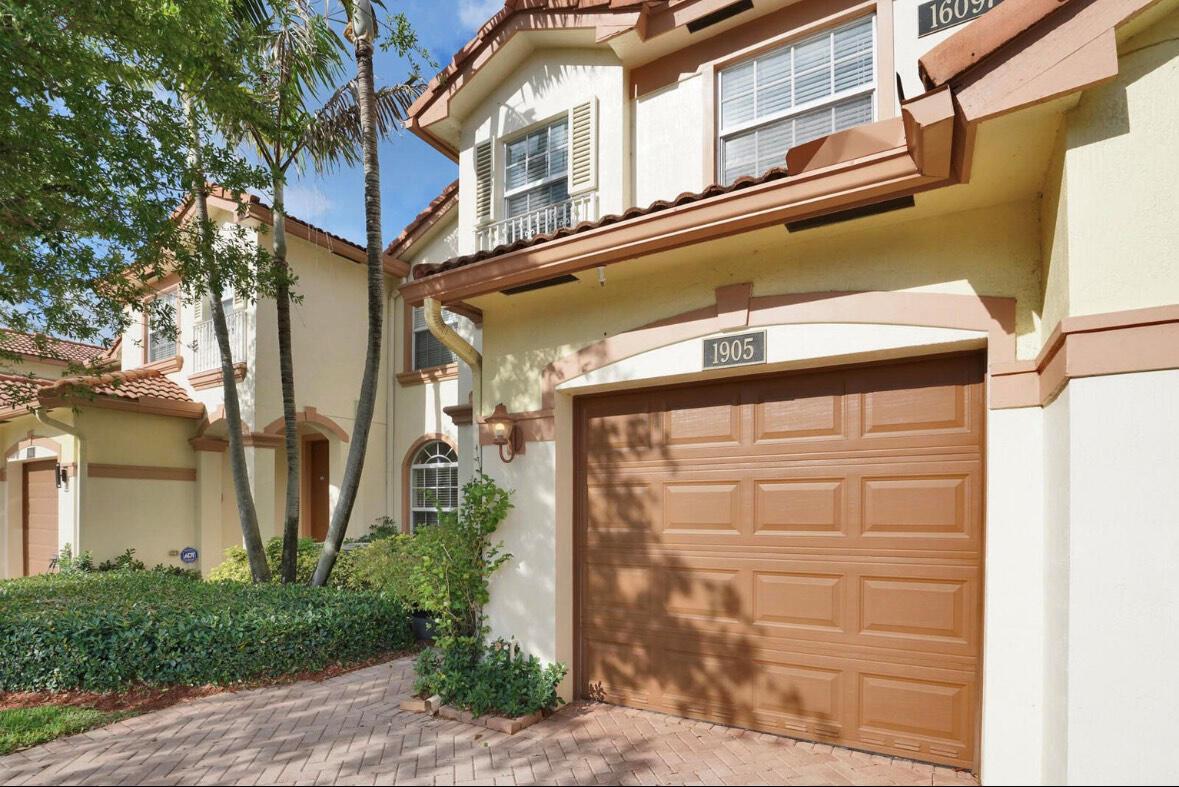 A two-story residential building features a brown garage door, beige stucco walls, and palm trees — 16097 Poppyseed Circle # 1905, Delray Beach FL 33484, DELRAY BEACH, FL