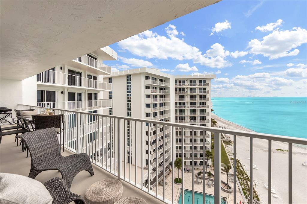 A balcony with wicker furniture overlooks a beach, swimming pool, and neighboring high-rise buildings — 2301 GULF OF MEXICO DRIVE UNIT 102N, LONGBOAT KEY, FL
