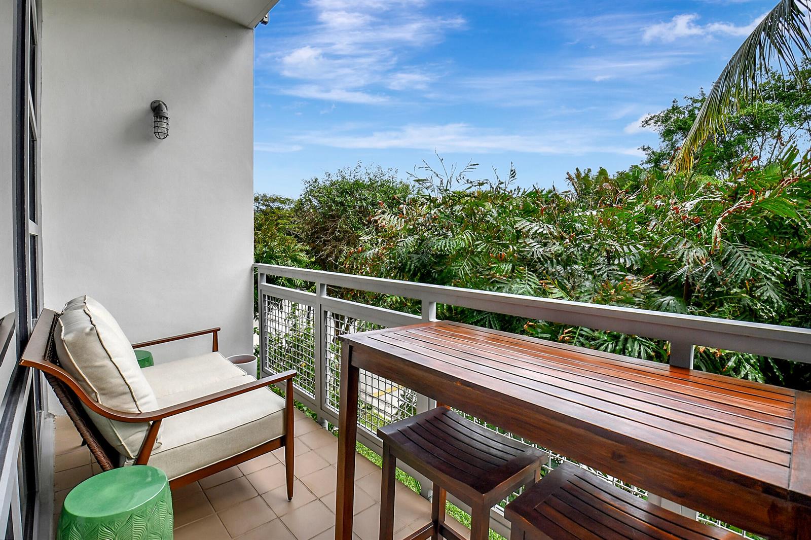 A balcony with a wooden table, stools, and a cushioned chair overlooking lush green trees — 365 SE 6th Avenue # 204, Delray Beach FL 33483, DELRAY BEACH, FL