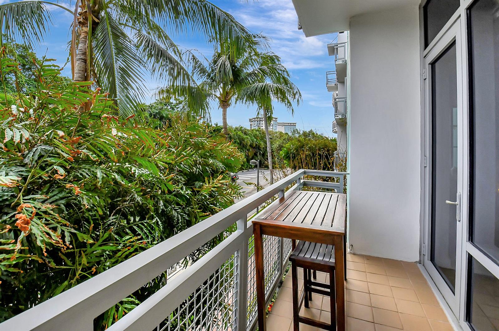 A balcony with a wooden table and stool overlooks a street lined with palm trees — 365 SE 6th Avenue # 204, Delray Beach FL 33483, DELRAY BEACH, FL