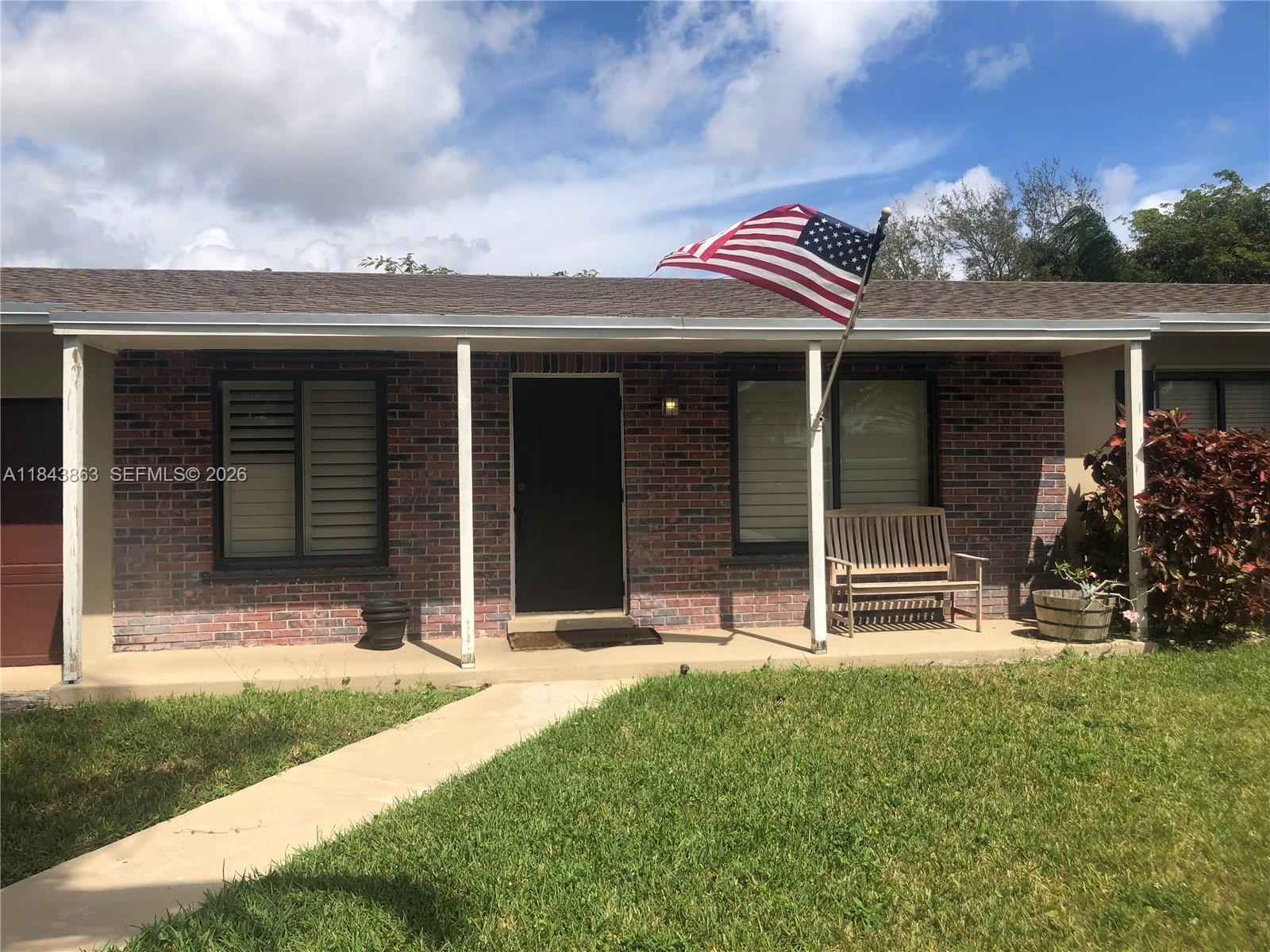 A single-story brick house features a covered porch, a wooden bench, and an American flag — 10801 SW 123rd St, Miami FL 33176, MIAMI, FL