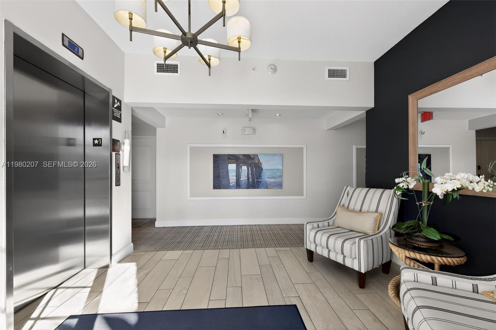 A modern elevator lobby features a striped armchair, a chandelier, and a framed pier painting — 915 S Bay Colony Dr S, Juno Beach FL 33408, JUNO BEACH, FL