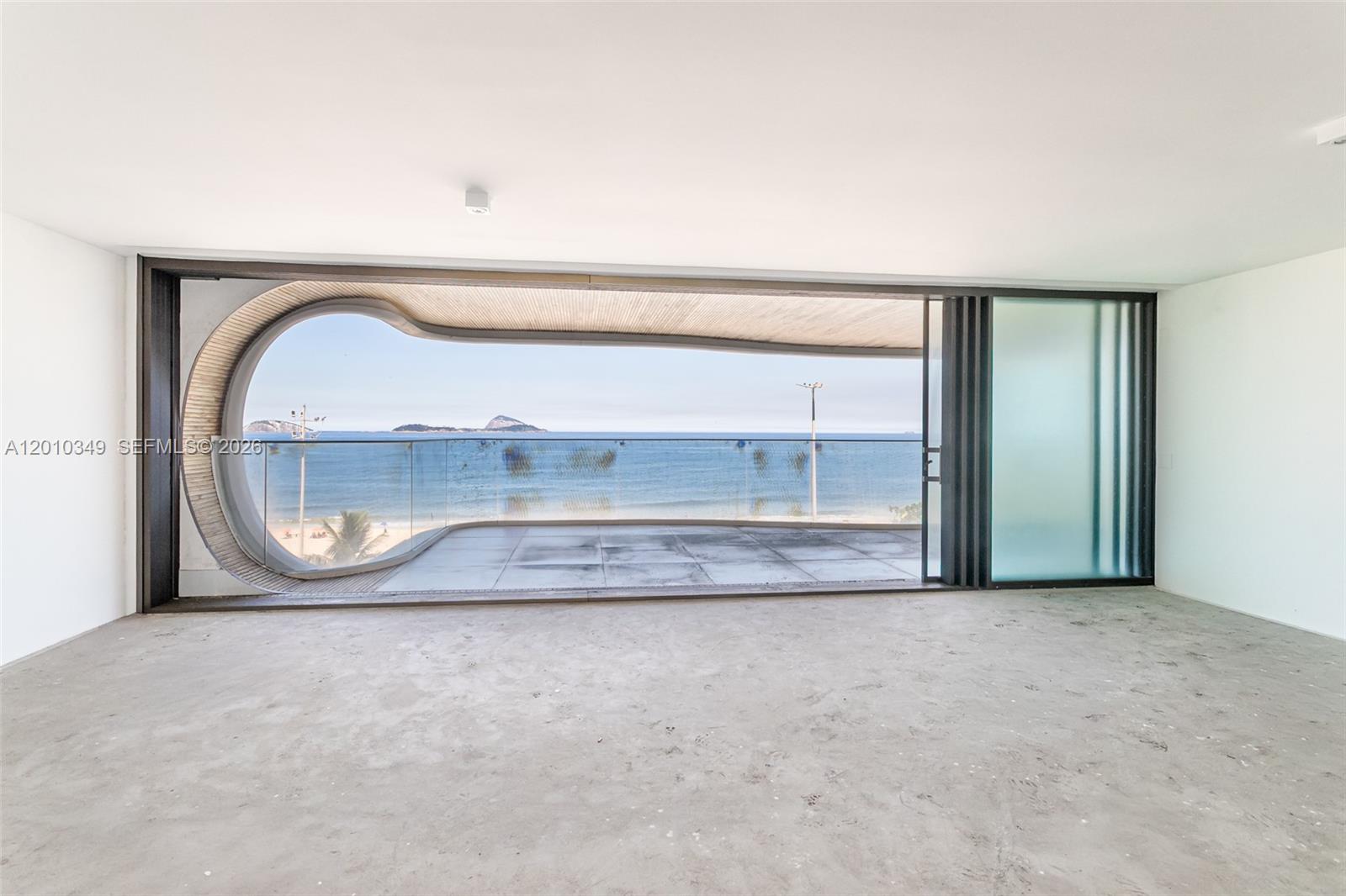 An unfurnished room features a large, uniquely curved window opening onto a balcony overlooking the ocean — FL