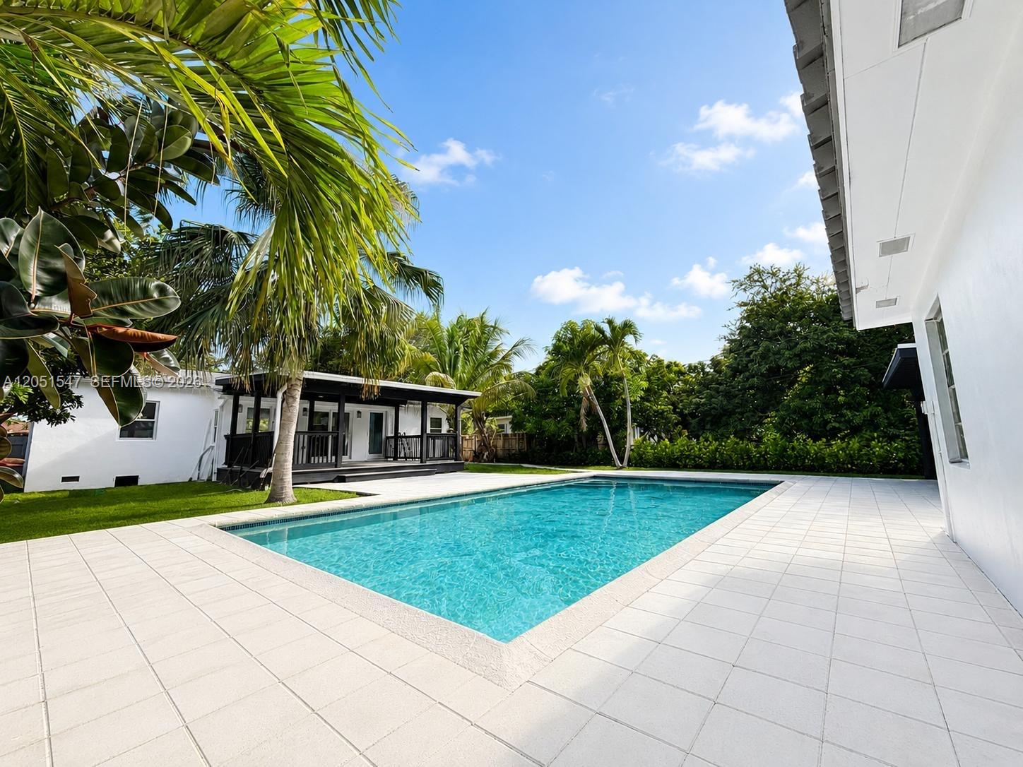 A rectangular swimming pool sits in a backyard patio area next to a white house — 859 NE 115th St, Biscayne Park FL 33161, BISCAYNE PARK, FL