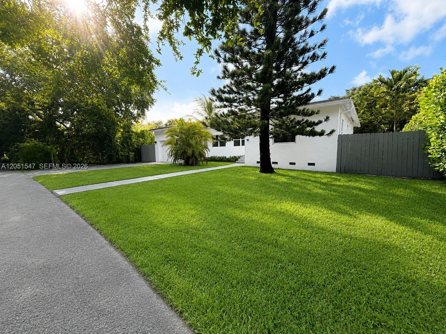 A white single-story house features a large front lawn, a pine tree, and gray fencing — 859 NE 115th St, Biscayne Park FL 33161, BISCAYNE PARK, FL