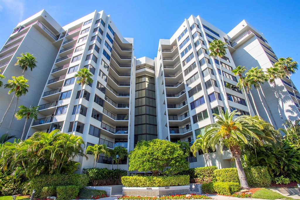 A low-angle exterior view of a multi-story white apartment building with balconies and palm trees — 1600 GULF BOULEVARD UNIT 512, CLEARWATER BEACH, FL